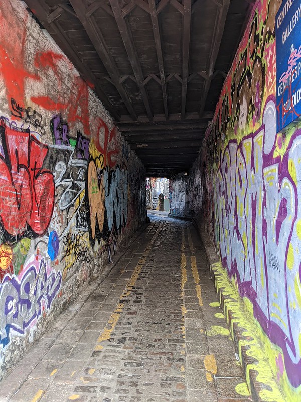 Graffitied walls on the side of a covered passage leading to the a back alley.