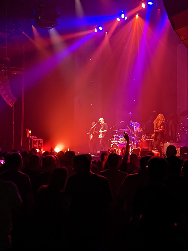 Devin Townsend on stage with his band against a dark red backdrop with shaded fans in the foreground