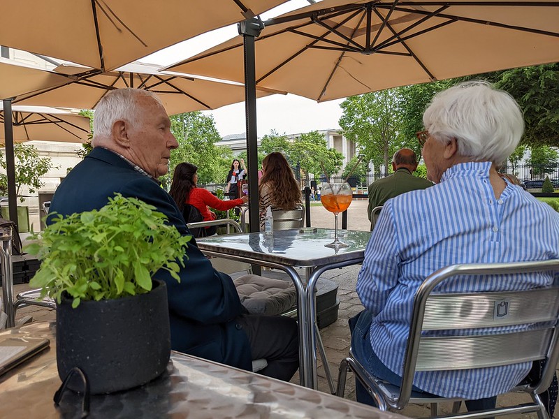 Elderly couple sitting at a table in a garden café sharing a cocktail with two straws.