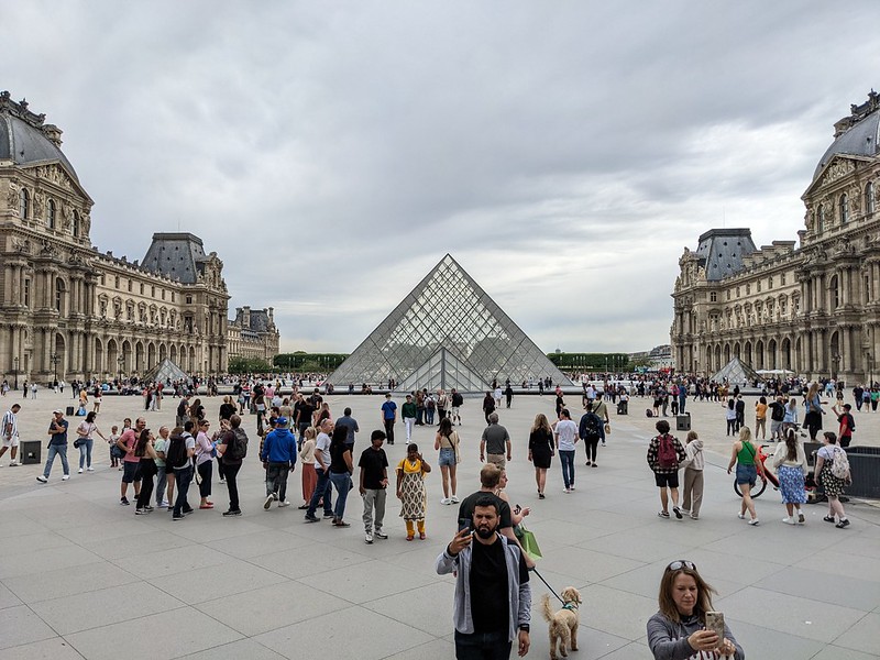 People milling around in the square in front of the Louvre with the glass pyramid in the background.