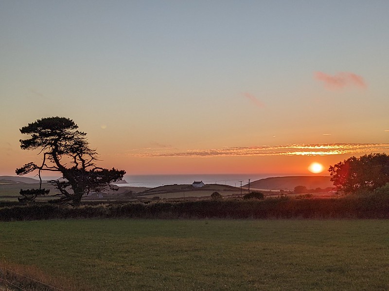 Sunsetting into the sea with a house on a hill just before the sea and fields in the foreground