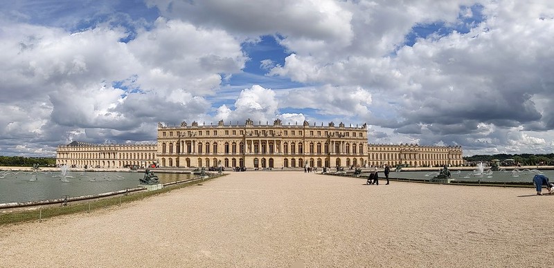 Panoramic view of Versailles castle in the distance with fountain pools on either side in the foreground and white clouds and blue sky above.