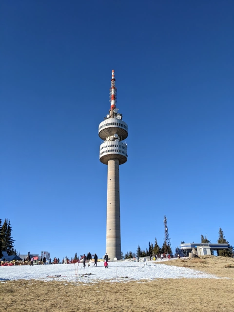 View of the radio tower at Snejanka, with blue sky background and brown fields in the foreground, except for the ski piste