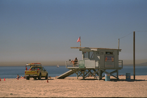 Lifeguard sitting in front on the porch of his watch tower, listening ot the radio, watching the sea, with his yellow jeep parked next to the tower